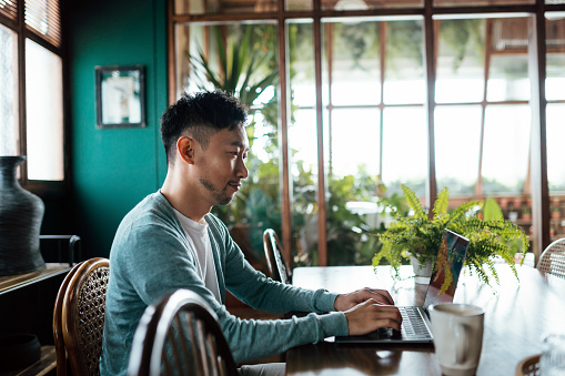 Young Asian man sitting at table, managing his finances and investment with online banking on laptop computer in living room at home. Technology makes life much easier