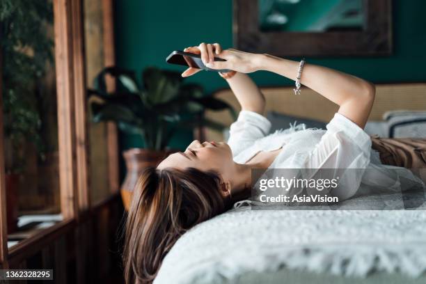 relaxed young asian woman checking social media on smartphone while lying in bed. youth culture. technology in everyday life - dating app stock pictures, royalty-free photos & images