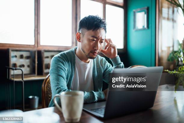 worried young asian man with his hand on head, using laptop computer at home, looking concerned and stressed out - press conference bildbanksfoton och bilder