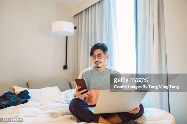 young handsome man using his smartphone while working from his laptop on bed during pandemic quarantine in hotel room - young man to bed photos et images de collection