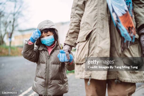 lovely little girl in protective face mask & warm clothing walking in town street with her mom joyfully - krankheitsverhinderung stock-fotos und bilder