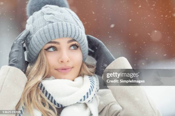 beautiful smiling blone girl fixes her knitted winter cap as she starts walking outdoors on a snowy cold day. - pop up store stock pictures, royalty-free photos & images