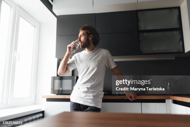 a handsome bearded guy stands and drinks water in the kitchen. modern cuisine. water. bearded guy. casual style. glass of water. - wasser trinken zu hause stock-fotos und bilder