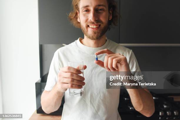 the guy drinks vitamins and pills in the kitchen. modern cuisine. water. young guy. casual style. glass of water. vitamins and dietary supplements. - allergy doctor stockfoto's en -beelden
