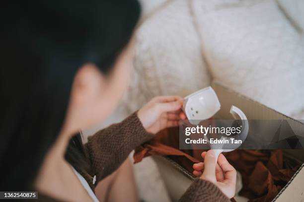 infeliz mujer china asiática sosteniendo tetera de cerámica rota de compras en línea en una sala de estar - devolución del saque fotografías e imágenes de stock