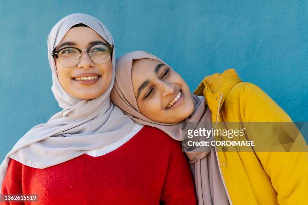 two young arab friends standing close to each other and smiling while posing outdoors. - arabische frau kopftuch stock-fotos und bilder