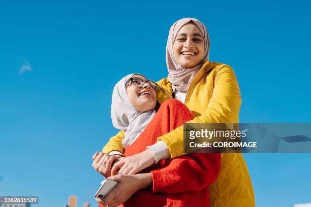 two muslim female friends hugging and smiling while posing outdoors against a clear blue sky. - islam foto e immagini stock