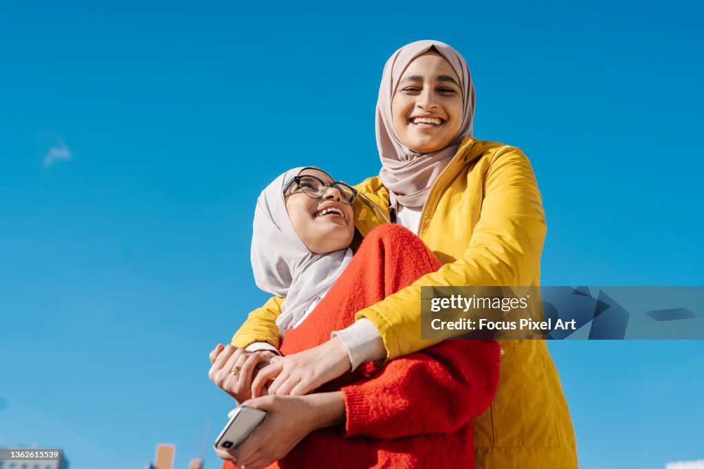 Two muslim female friends hugging and smiling while posing outdoors against a clear blue sky.