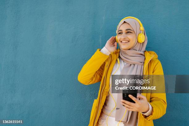 arab woman enjoying listening music with a smartphone and headphones while standing outdoors on the street. - middle east friends stock pictures, royalty-free photos & images