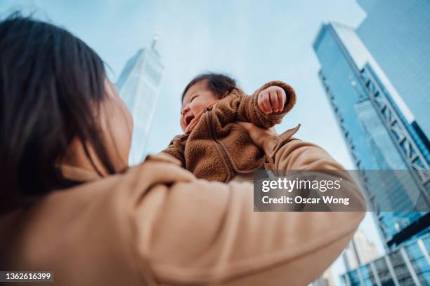 over the shoulder view of young asian mother lifting cheerful baby girl up in the air against modern skyscrapers. - over the shoulder view stock-fotos und bilder