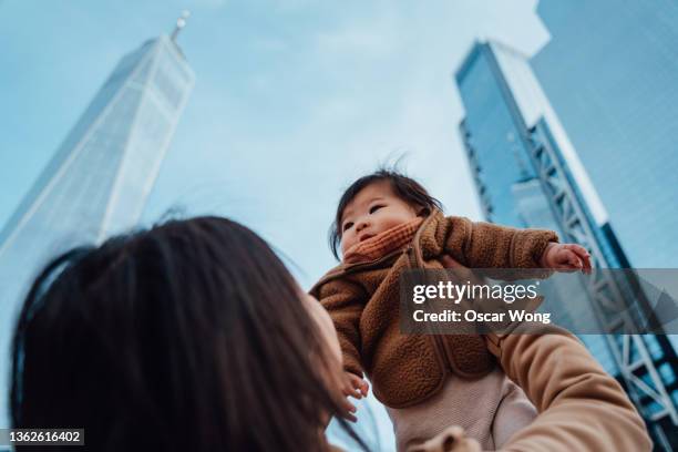 over the shoulder view of young asian mother lifting cheerful baby girl up in the air against modern skyscrapers. - over the shoulder view stock-fotos und bilder