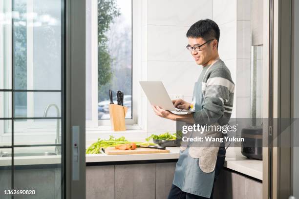 frontal photo of a young man standing in the kitchen using a computer - laptop frontal stock pictures, royalty-free photos & images