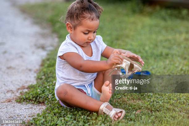 kleines mädchen, das auf dem gras sitzt und ihre sandale anzieht - girls wearing sandals stock-fotos und bilder