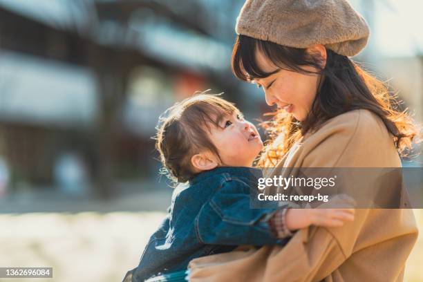 mother and small girl sitting at bench and enjoying spending time together in city in winter on sunny warm day - asian mum stockfoto's en -beelden
