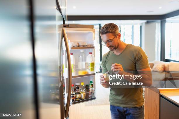 man eating yogurt at open refrigerator in kitchen - open fridge stock-fotos und bilder