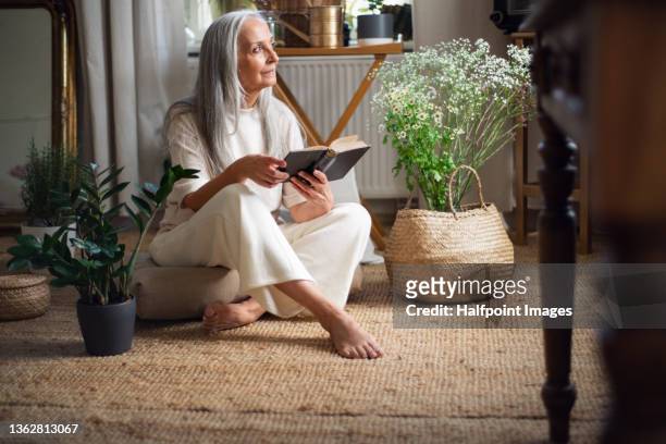 relaxed senior woman sitting on floor and reading book indoors at home. - texto religioso - fotografias e filmes do acervo