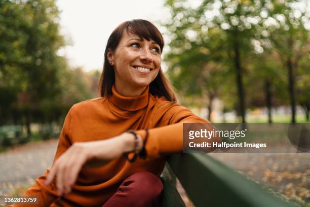 beautiful mid adult woman sitting on bench and resting outside in the city on autumn day. - women happy photos et images de collection