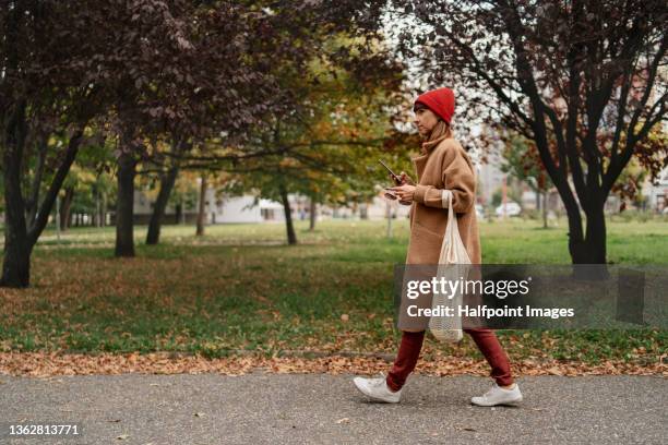 mid adult woman with mobile phone and reusable shopping bag walking outside in the city on autumn day. - casacca foto e immagini stock