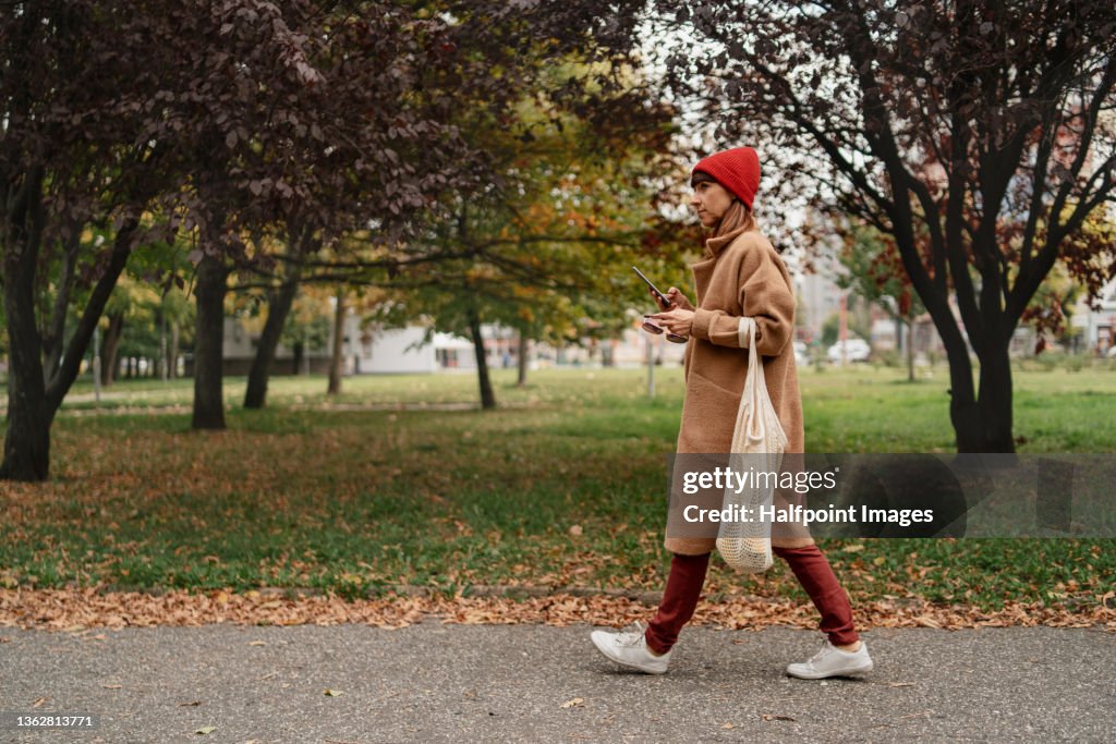 Mid adult woman with mobile phone and reusable shopping bag walking outside in the city on autumn day.