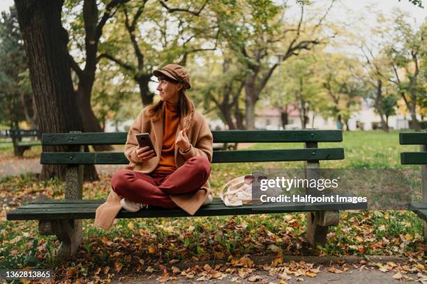 beautiful mid adult woman sitting on bench and using mobile phone outside in the city on autumn day. - banco del parque fotografías e imágenes de stock