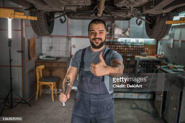 gutaussehender bärtiger mechaniker, der in der autowerkstatt steht und ein erhöhtes daumenzeichen zeigt - daumen stock-fotos und bilder