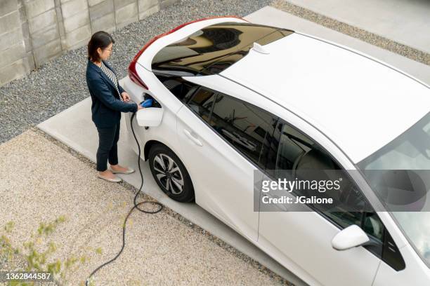 mittelhohe frau, die ihr elektroauto zu hause auflädt - chargers stock-fotos und bilder