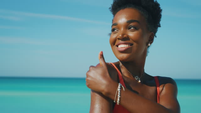 행복한 젊은 아프리카 여성의 슬로우 모션은 해변에서 휴가를 보내고 카메라에서 미소짓는 동안 피부를 돌보기 위해 자외선 차단제 또는 선 탠딩 로션을 바르고 있습니다.