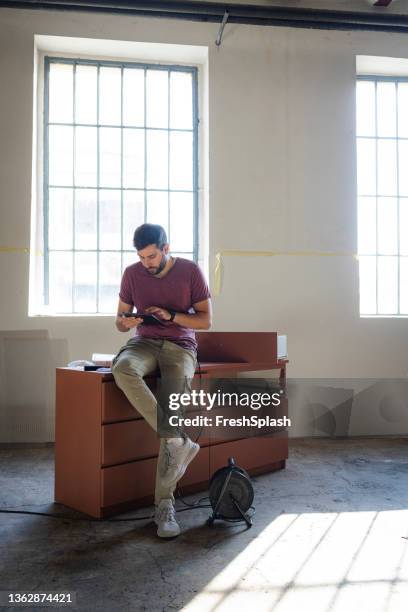 young furniture designer sitting on a wooden chest of drawers and using a digital tablet for work - dresser stock pictures, royalty-free photos & images