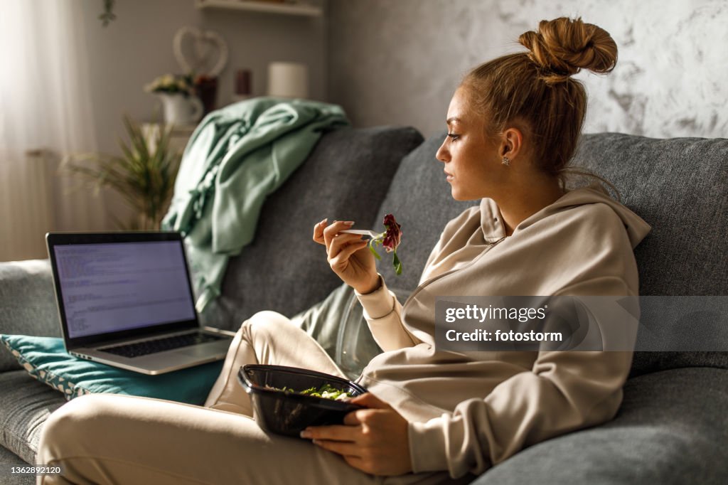 Adolescente sentada en el sofá, estudiando y comiendo una ensalada para llevar