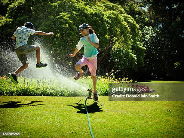 sibling jumping over water sprinkler - sprinkler stock pictures, royalty-free photos & images