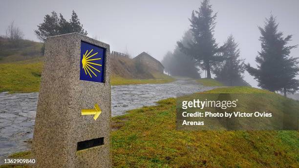 signpost of the saint james way displaying the traditional yellow arrow and scallop on a rainy foggy day in the village of o cebreiro - santiago de compostela stockfoto's en -beelden