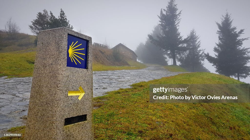 Signpost of the Saint James Way displaying the traditional yellow arrow and scallop on a rainy foggy day in the village of O Cebreiro