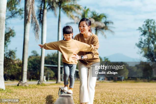 joyful young asian mother and little daughter spending time together in nature park on a sunny day. mother assisting little daughter while balancing on wooden plank with her arms outstretched. family lifestyle, love and care concept - trust stock pictures, royalty-free photos & images