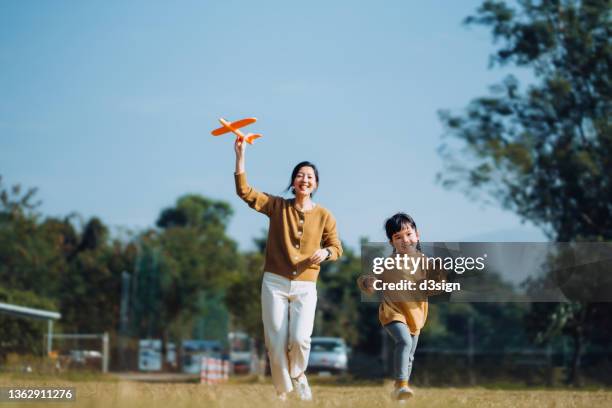 young asian mother and lovely little daughter spending time together outdoors, playing with airplane toy and smiling joyfully in park on a lovely sunny day against beautiful blue sky - mother running stockfoto's en -beelden