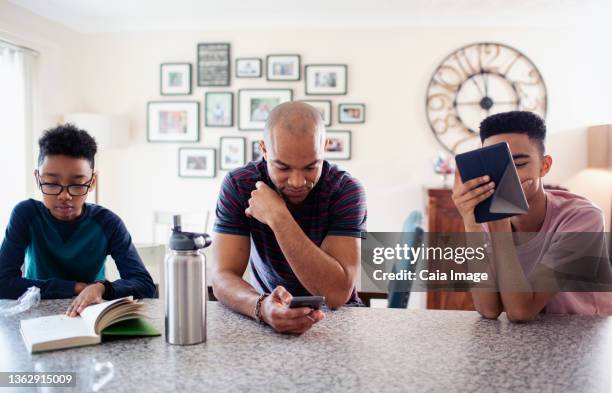 father and sons using digital tablet, smart phone and reading book in kitchen - father stock pictures, royalty-free photos & images