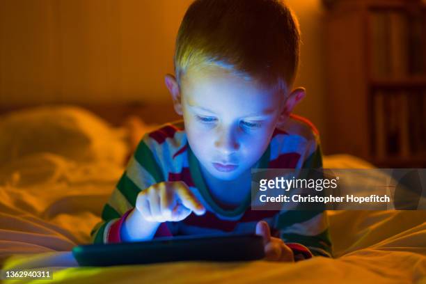 a young boy lying on his parent's bed concentrating on a tablet computer, his face illuminated by the screen - ipad glow foto e immagini stock