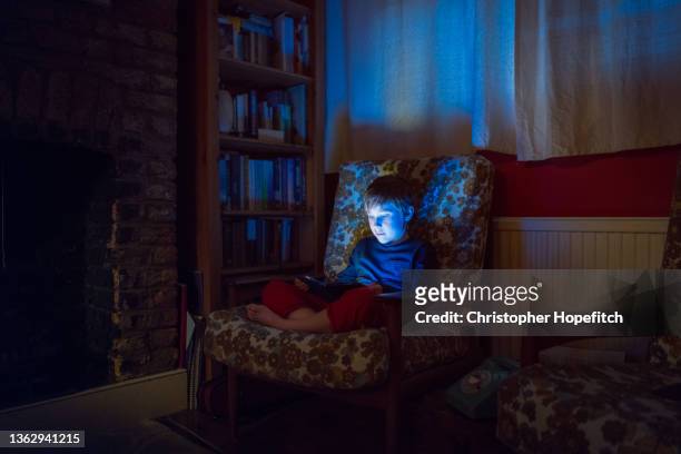 a young boy sat on a sofa in a living room at night, his face illuminated by the screen of the smartphone he is looking at. - children screen stock-fotos und bilder