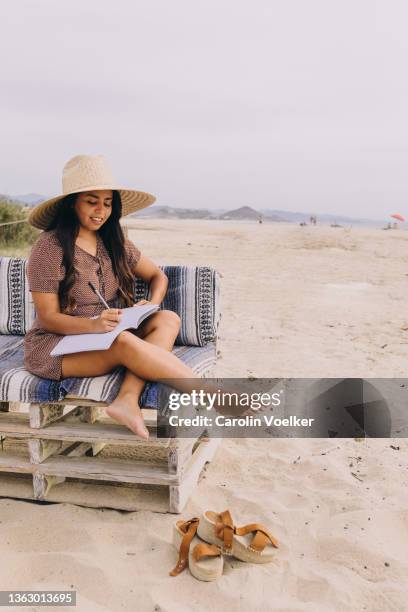latina young women on the beach with a hat sitting in an outdoor chair writing - gambe incrociate foto e immagini stock