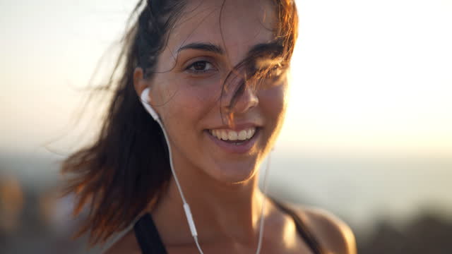An young active sporty athlete woman with firm fit body is smiling in camera satisfied with her achievement results after running and jogging workout on a top of rock with seascape at sunset.