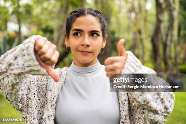 young latino woman looking at the camera confused and giving a thumbs up and thumbs down, pointing - marketing no advertising campaigns stock pictures, royalty-free photos & images