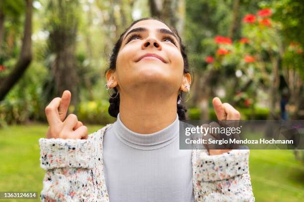 young latino woman looking up and crossing her fingers in hope, wishing - sorte imagens e fotografias de stock