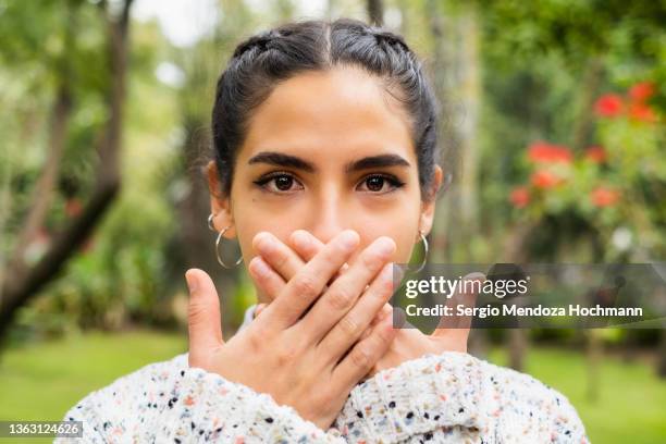 young latino woman looking at the camera and covering her mouth, speak no evil - people whispering imagens e fotografias de stock