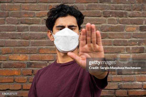 young latino man wearing a face mask, looking at the camera, and gesturing to stop - stop ストックフォトと画像