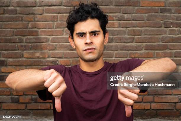 young latino man looking at the camera and giving a thumbs down - guy with attitude mid shot stock pictures, royalty-free photos & images