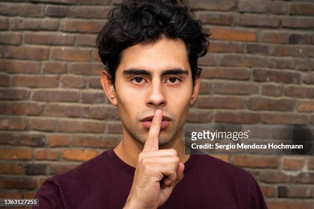 young latino man looking at the camera with a finger on his lips, asking for quiet, silence - finger in mouth stockfoto's en -beelden