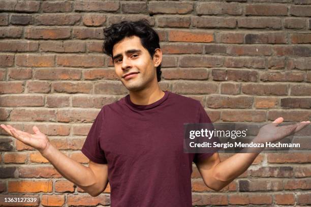 young latino man looking at the camera and shrugging with his arms outstretched - shrug shoulders stockfoto's en -beelden