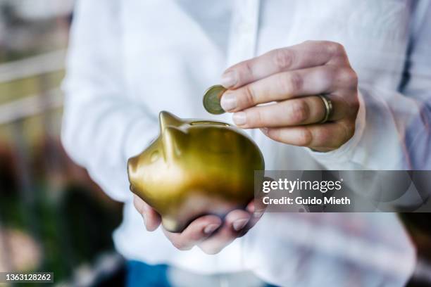 woman putting savings in a golden piggy bank while standing in front of a window. - gold investment stock pictures, royalty-free photos & images