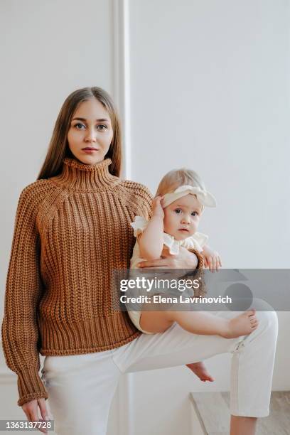 stylish mom and daughter are posing against the backdrop of a light wall. mothers day. portrait of a young woman with a one-year-old baby in her arms - fashionable mom stock pictures, royalty-free photos & images