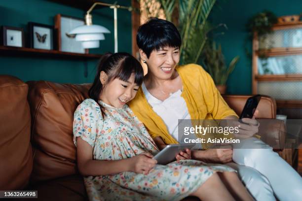 lächelnde asiatische großmutter und enkelin entspannen sich im wohnzimmer zu hause, großmutter mit smartphone, während enkelin auf digitales tablet schaut. technologieeinsatz über alle altersgruppen hinweg - family phone tablet stock-fotos und bilder