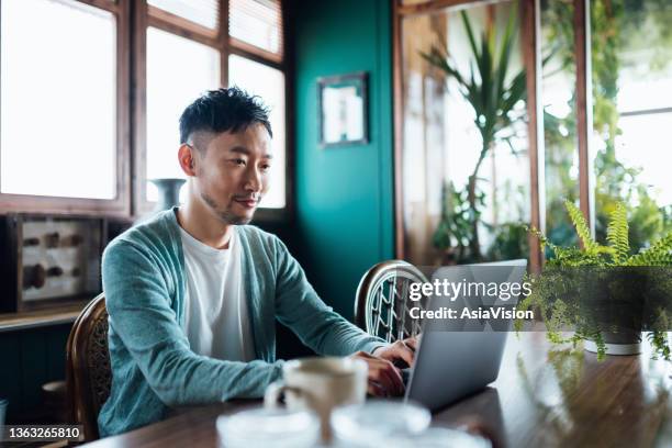 jeune homme asiatique souriant assis à la table de la salle à manger à la maison, faisant des achats en ligne avec un ordinateur portable. concept de paiement en ligne pratique et sûr. la technologie rend la vie beaucoup plus facile - self employed photos et images de collection